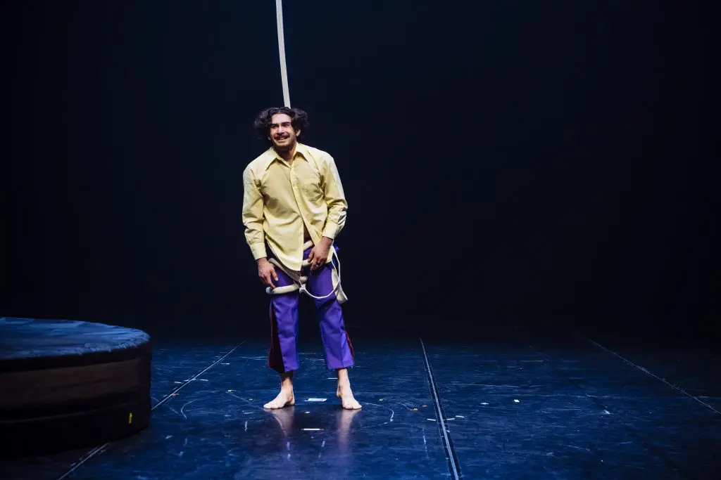 Dylan Ingwersen, pieds nus, cheveux aux épaules, vêtu d'une chemise jaune et d'un pantalon violet, se tient sur une scène sombre en tenant une corde suspendue, avec une plate-forme ronde rembourrée à proximité.