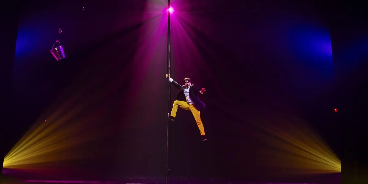 Antoine Brabant, vêtu d'un pantalon jaune et d'une veste de costume, grimpe à un grand mât sur une scène éclairée par des projecteurs, avec deux seaux métalliques suspendus au-dessus et des pétales éparpillés sur le sol, le tout sur un fond sombre et dramatique.