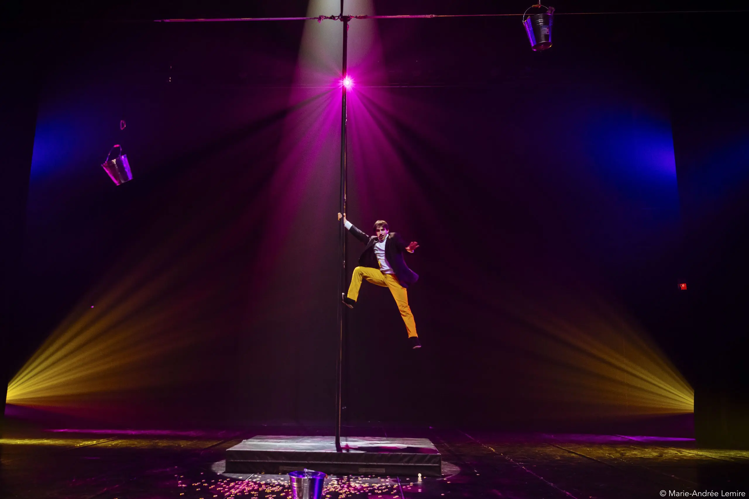 Antoine Brabant, vêtu d'un pantalon jaune et d'une veste de costume, grimpe à un grand mât sur une scène éclairée par des projecteurs, avec deux seaux métalliques suspendus au-dessus et des pétales éparpillés sur le sol, le tout sur un fond sombre et dramatique.
