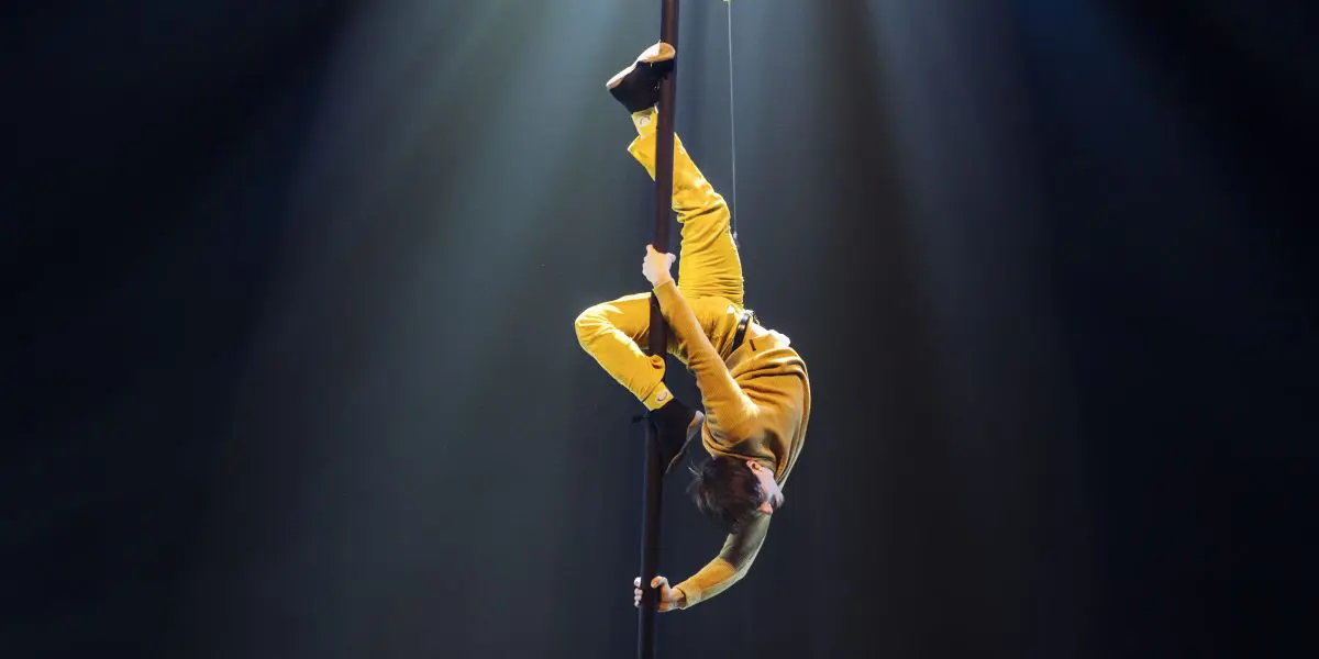Antoine Brabant, vêtu d'une tenue jaune, exécute une pose acrobatique sur un poteau vertical, éclairé par un éclairage de scène dramatique sur un fond sombre.