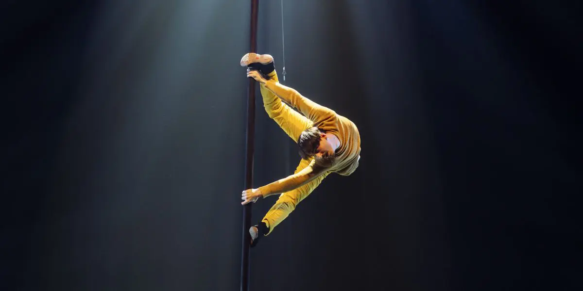Un artiste en tenue jaune se tient en équilibre horizontal sur un poteau vertical, les jambes écartées et les bras agrippés au poteau. Antoine Brabant commande la scène, éclairée par un éclairage dramatique sur un fond sombre.