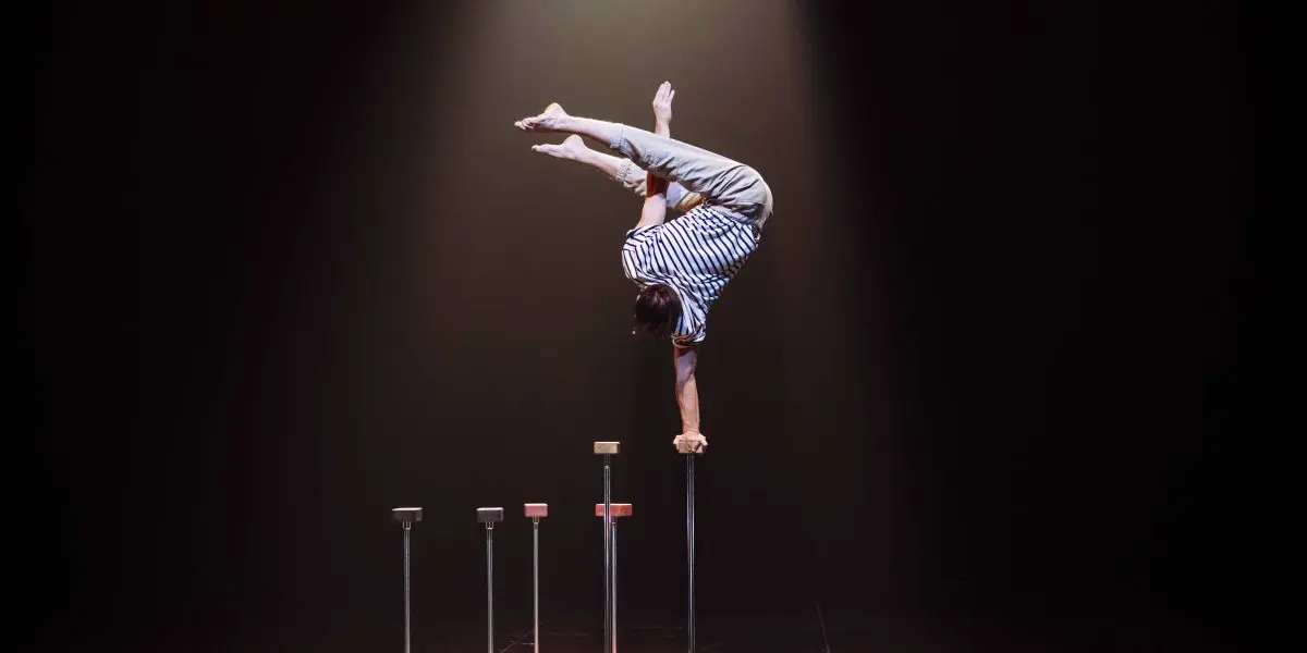 Sur une scène faiblement éclairée, Carl Löwenborg se tient en équilibre, la tête en bas, sur une main, au sommet d'un grand bâton, les jambes pliées. Quatre autres bâtons de différentes hauteurs sont alignés à côté de lui, soulignant son habileté et sa précision remarquables.