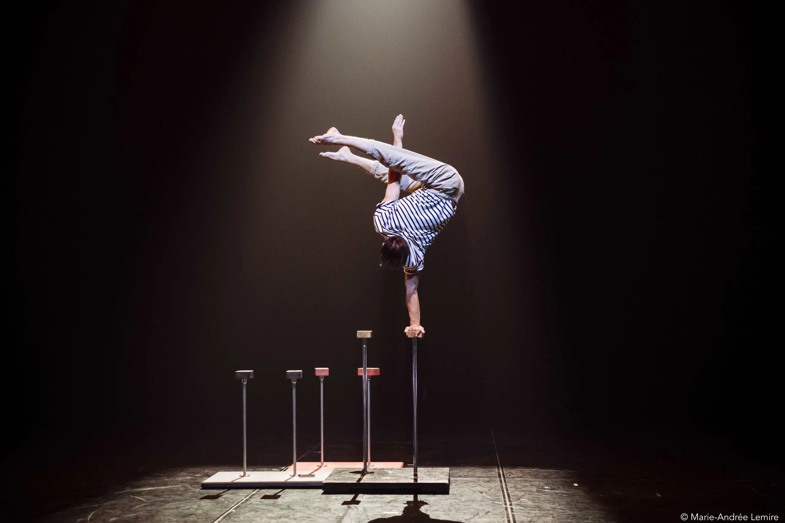 Sur une scène faiblement éclairée, Carl Löwenborg se tient en équilibre, la tête en bas, sur une main, au sommet d'un grand bâton, les jambes pliées. Quatre autres bâtons de différentes hauteurs sont alignés à côté de lui, soulignant son habileté et sa précision remarquables.