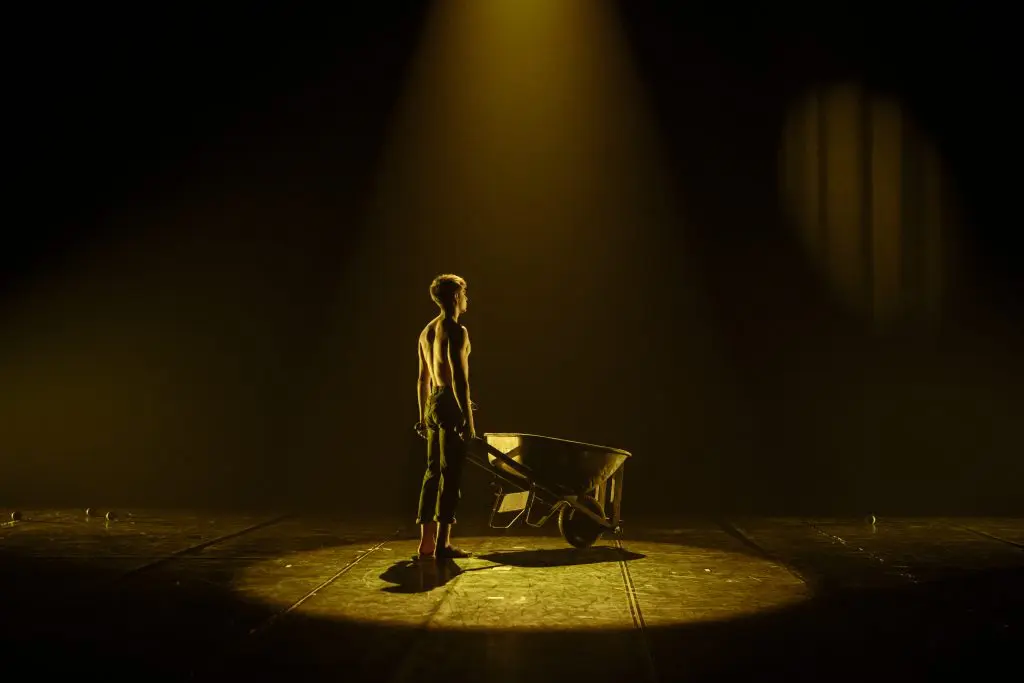 Une personne torse nu se tient sur une scène faiblement éclairée, tenant les poignées d'une brouette vide. Un projecteur unique les éclaire, projetant des ombres dramatiques sur le sol - une scène évocatrice qui rappelle les performances expressives de Basile Pucek.