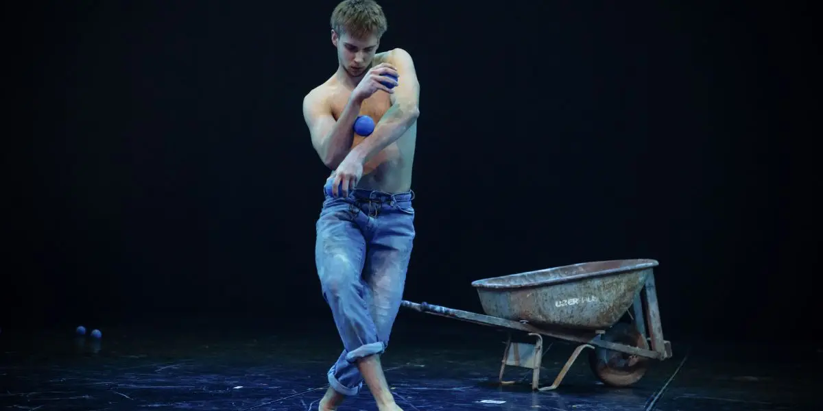 Basile Pucek, pieds nus et torse nu en jeans, jongle avec des balles bleues sur une scène sombre avec une brouette en arrière-plan. De la poudre blanche recouvre son corps et son jean, et d'autres balles sont éparpillées sur le sol.