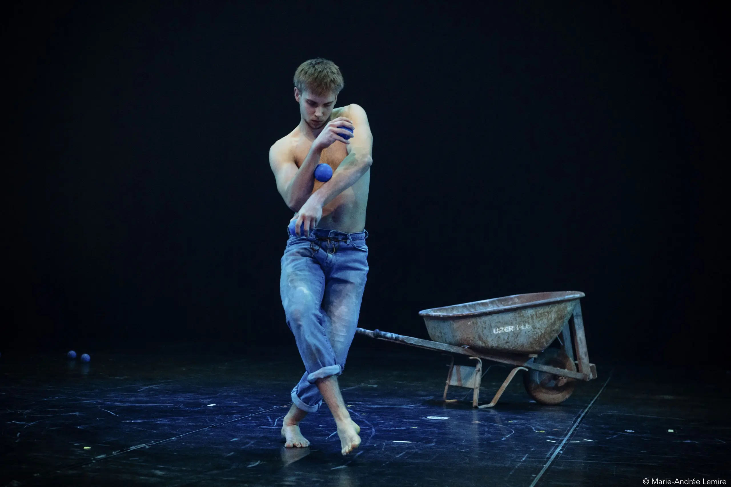 Basile Pucek, pieds nus et torse nu en jeans, jongle avec des balles bleues sur une scène sombre avec une brouette en arrière-plan. De la poudre blanche recouvre son corps et son jean, et d'autres balles sont éparpillées sur le sol.