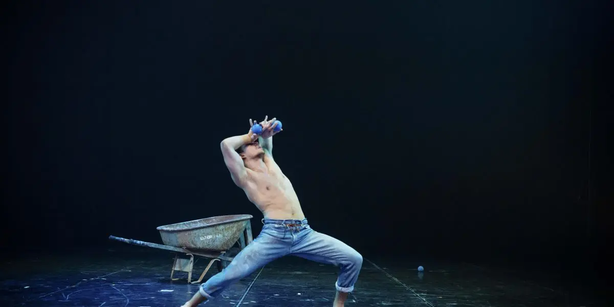 Une personne torse nu en jeans prend une pose de danse dramatique sur scène, penchée en arrière, les bras levés, à côté d'une brouette sous un éclairage théâtral - un moment évocateur d'un spectacle de Basile Pucek.