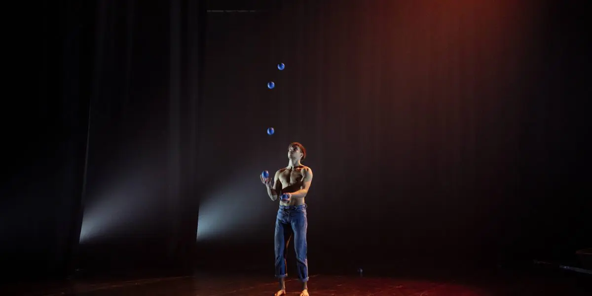 Une personne en jean et débardeur jongle avec cinq balles bleues sur une scène sombre à l'éclairage dramatique, créant une atmosphère concentrée et artistique qui rappelle les performances captivantes de Basile Pucek.