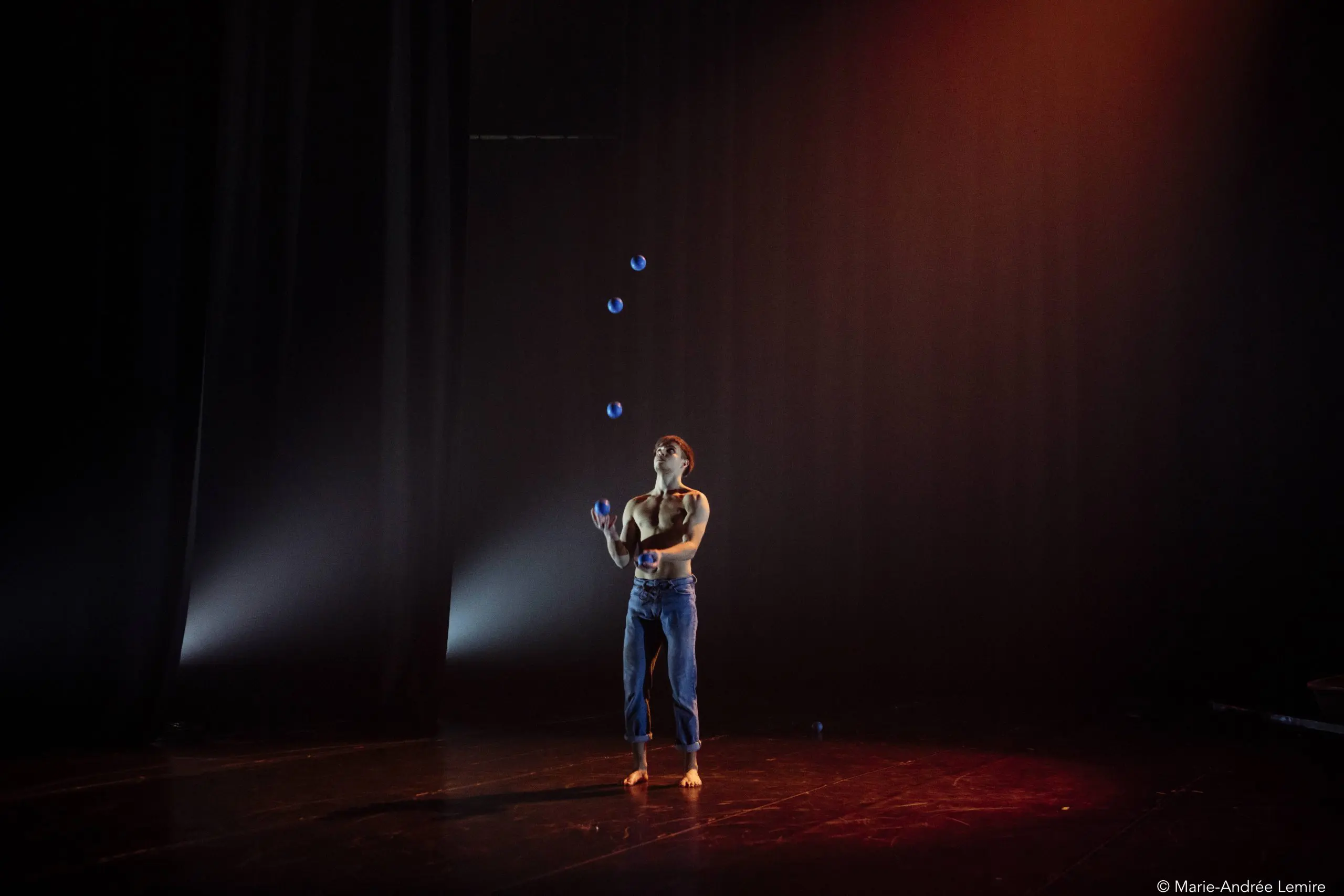Une personne en jean et débardeur jongle avec cinq balles bleues sur une scène sombre à l'éclairage dramatique, créant une atmosphère concentrée et artistique qui rappelle les performances captivantes de Basile Pucek.