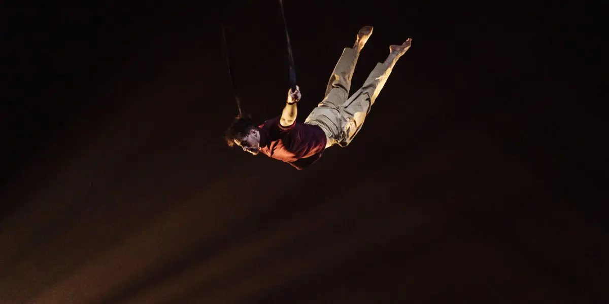 Eduardo De Azevedo Grillo, vêtu d'une chemise rouge et d'un pantalon beige, exécute un numéro aérien, suspendu à l'envers par des sangles, dans un décor sombre et éclairé par des projecteurs.