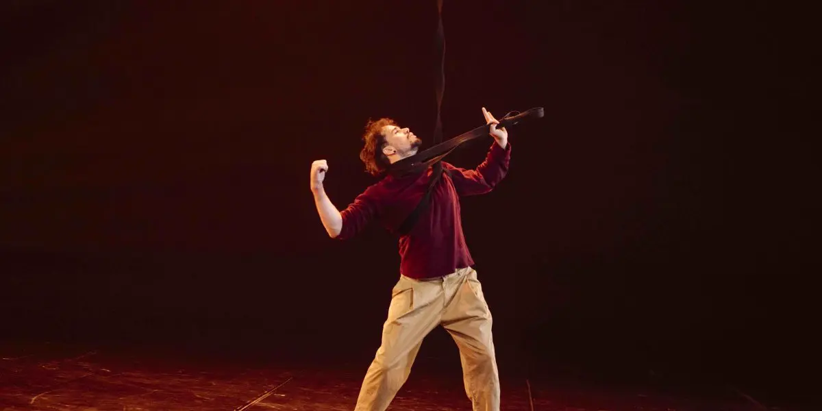 Une personne vêtue d'une chemise marron et d'un pantalon beige se tient pieds nus sur une scène faiblement éclairée, penchée en arrière, les bras repliés, tandis qu'un ruban ou une sangle noire est tendu sur ses épaules et son cou, évoquant le style de performance intense d'Eduardo De Azevedo Grillo.