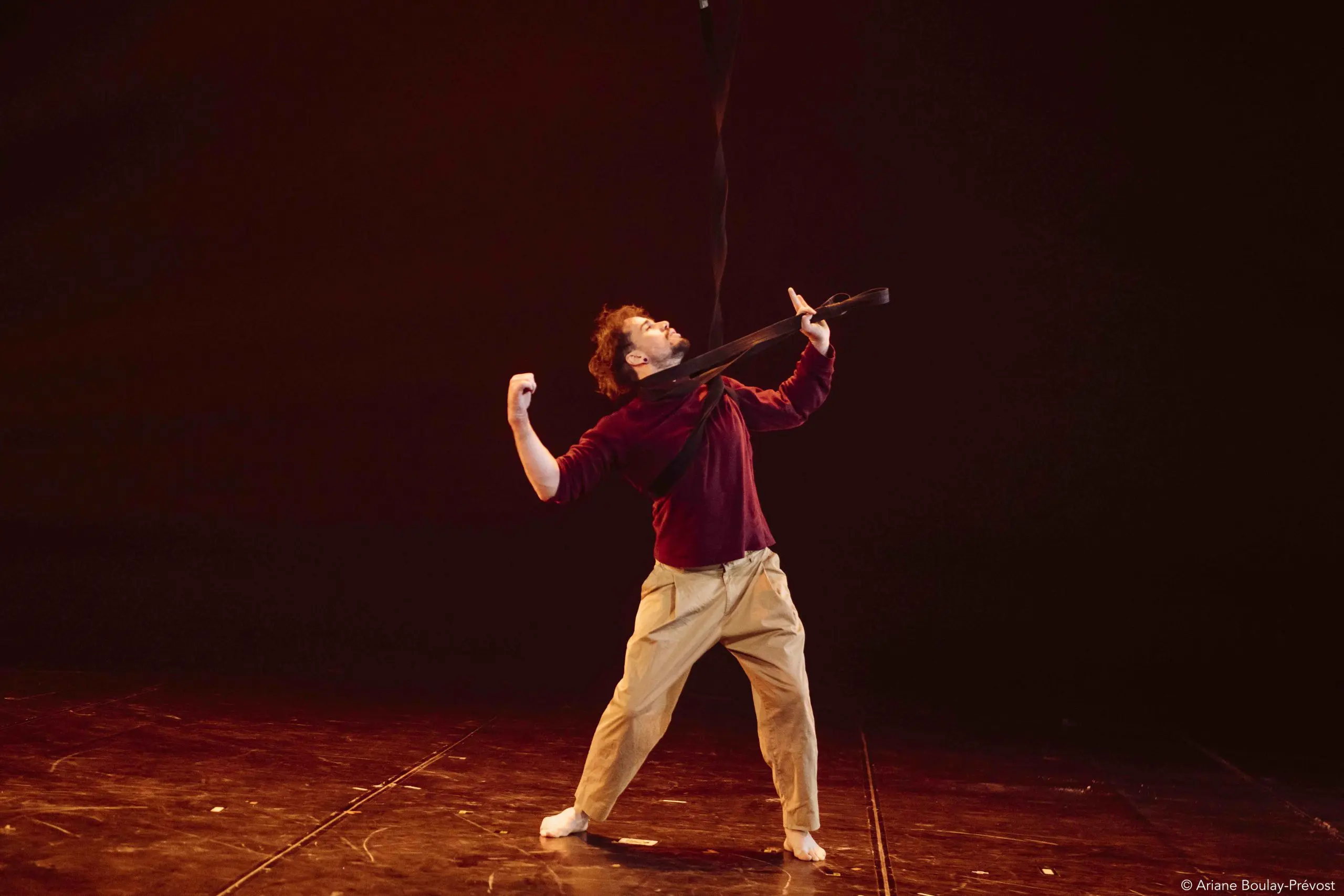 Une personne vêtue d'une chemise marron et d'un pantalon beige se tient pieds nus sur une scène faiblement éclairée, penchée en arrière, les bras repliés, tandis qu'un ruban ou une sangle noire est tendu sur ses épaules et son cou, évoquant le style de performance intense d'Eduardo De Azevedo Grillo.