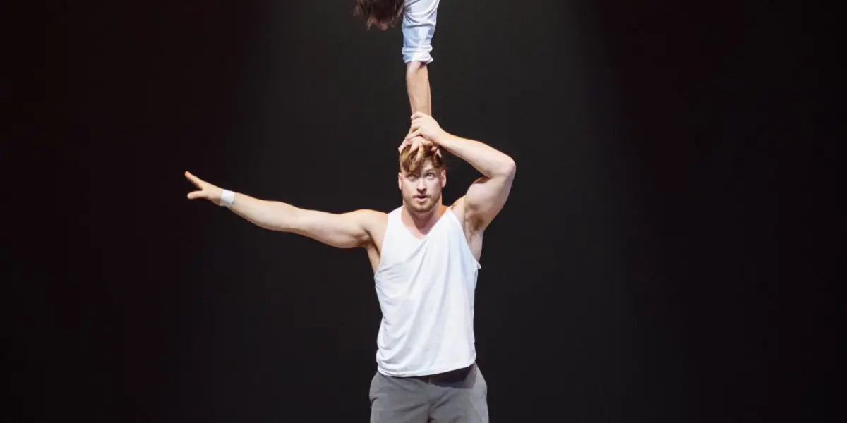 Deux acrobates masculins se produisent sur scène ; Carl Löwenborg se tient debout sur un tapis, l'air grave, et soutient l'autre, qui est en équilibre sur une main au-dessus de sa tête, les jambes étendues en fente sous un projecteur.