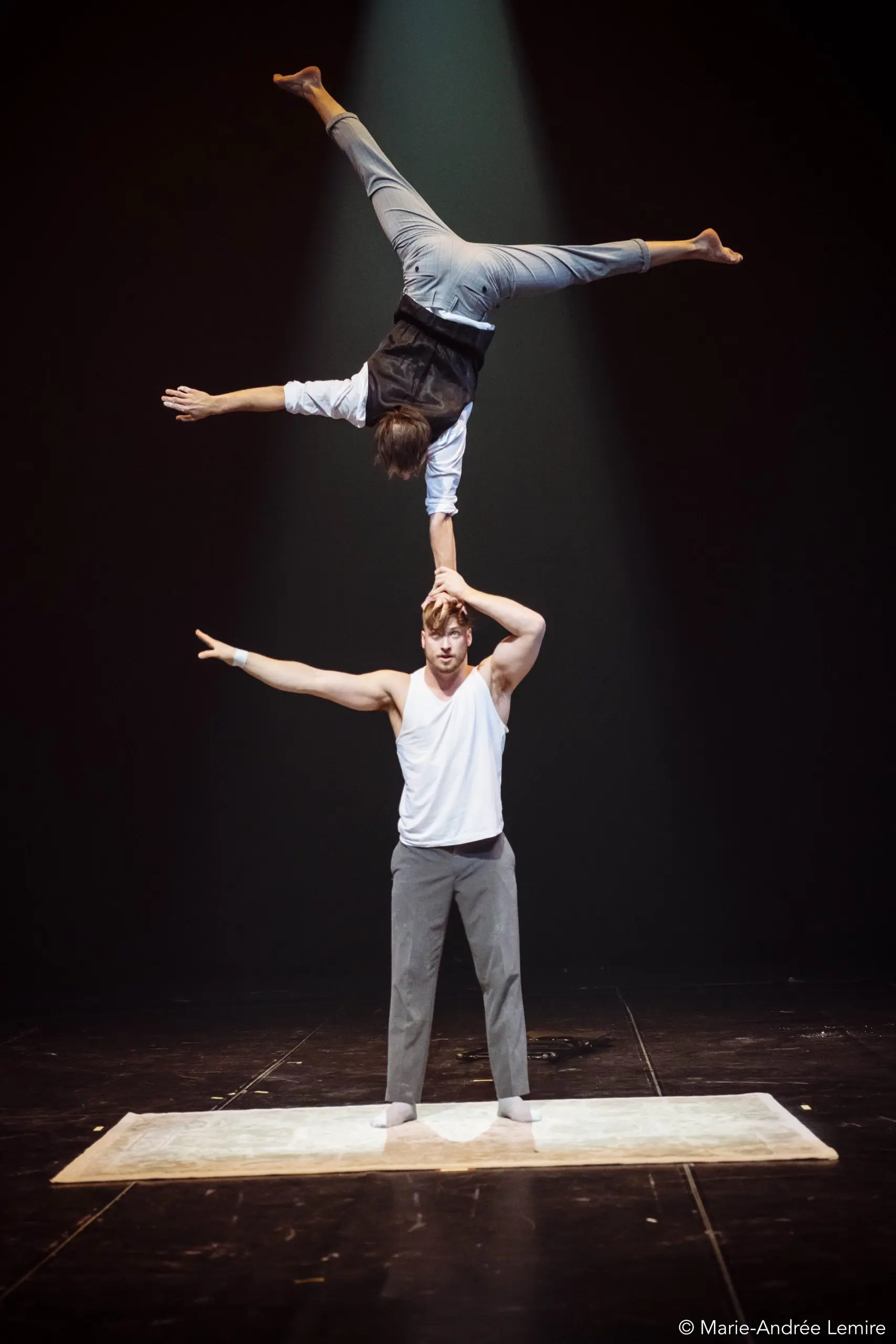 Deux acrobates masculins se produisent sur scène ; Carl Löwenborg se tient debout sur un tapis, l'air grave, et soutient l'autre, qui est en équilibre sur une main au-dessus de sa tête, les jambes étendues en fente sous un projecteur.
