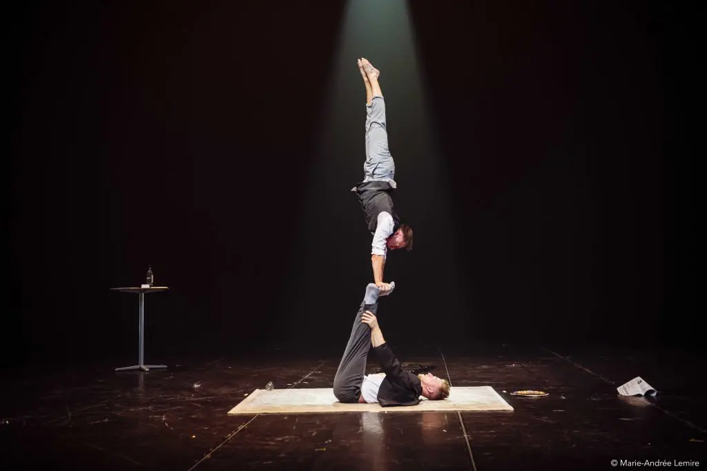 Deux artistes, dont Carl Löwenborg, sont en équilibre sur scène ; l'un est allongé sur le dos et tient l'autre verticalement en équilibre sur les mains. La scène est faiblement éclairée, avec une table et une bouteille en arrière-plan.