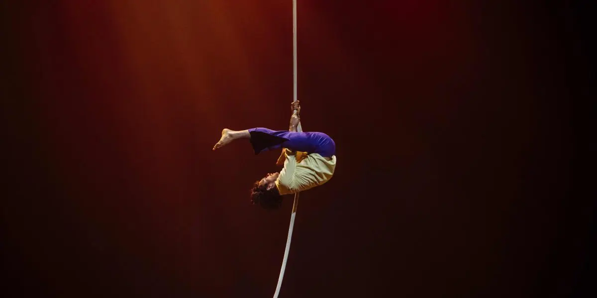 Dylan Ingwersen, vêtu d'une chemise jaune et d'un pantalon bleu, effectue une pose acrobatique, suspendu à l'envers à une corde blanche sur un fond sombre et chaudement éclairé.