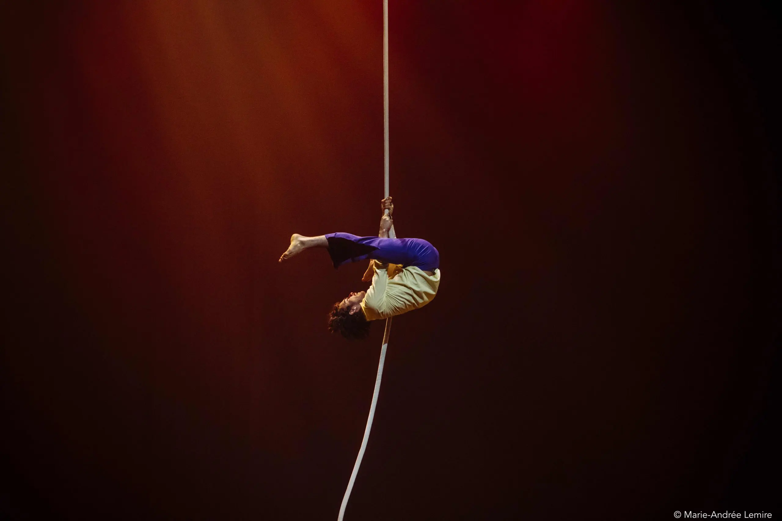 Dylan Ingwersen, vêtu d'une chemise jaune et d'un pantalon bleu, effectue une pose acrobatique, suspendu à l'envers à une corde blanche sur un fond sombre et chaudement éclairé.