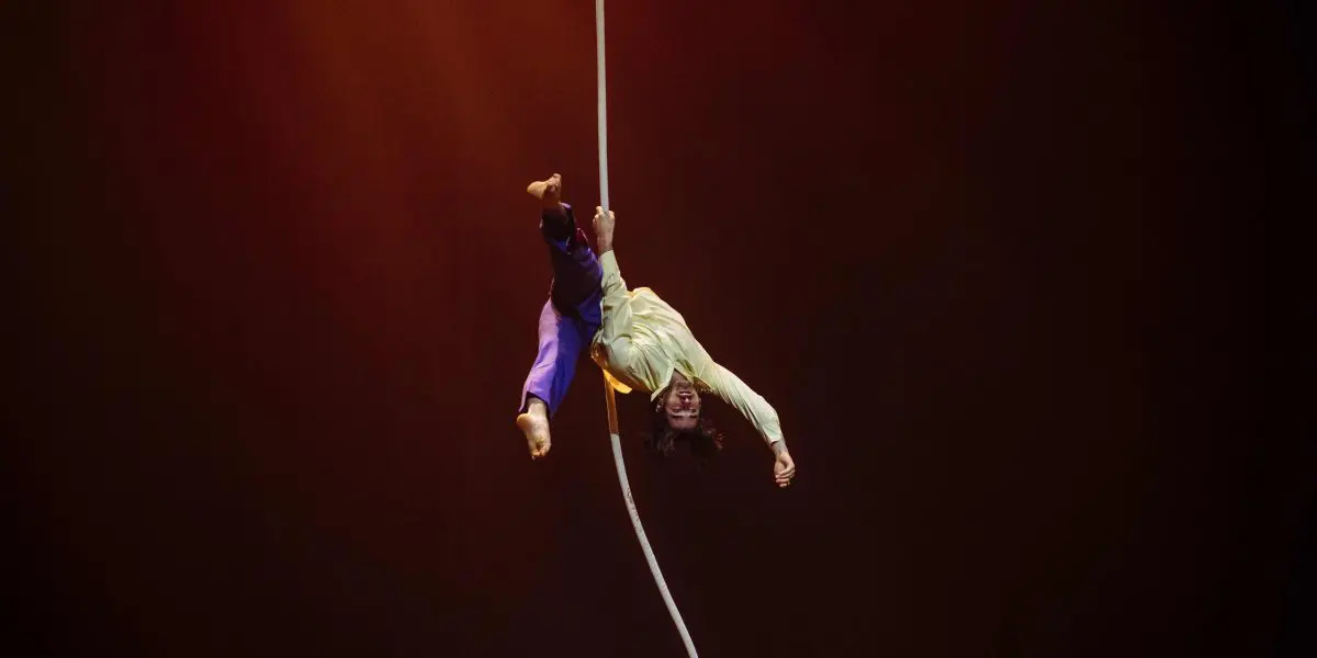 Un artiste de cirque, Dylan Ingwersen, est suspendu à l'envers à une corde aérienne blanche, en plein acte, un bras tendu et les jambes pliées, sur un fond de scène sombre à l'éclairage chaud.