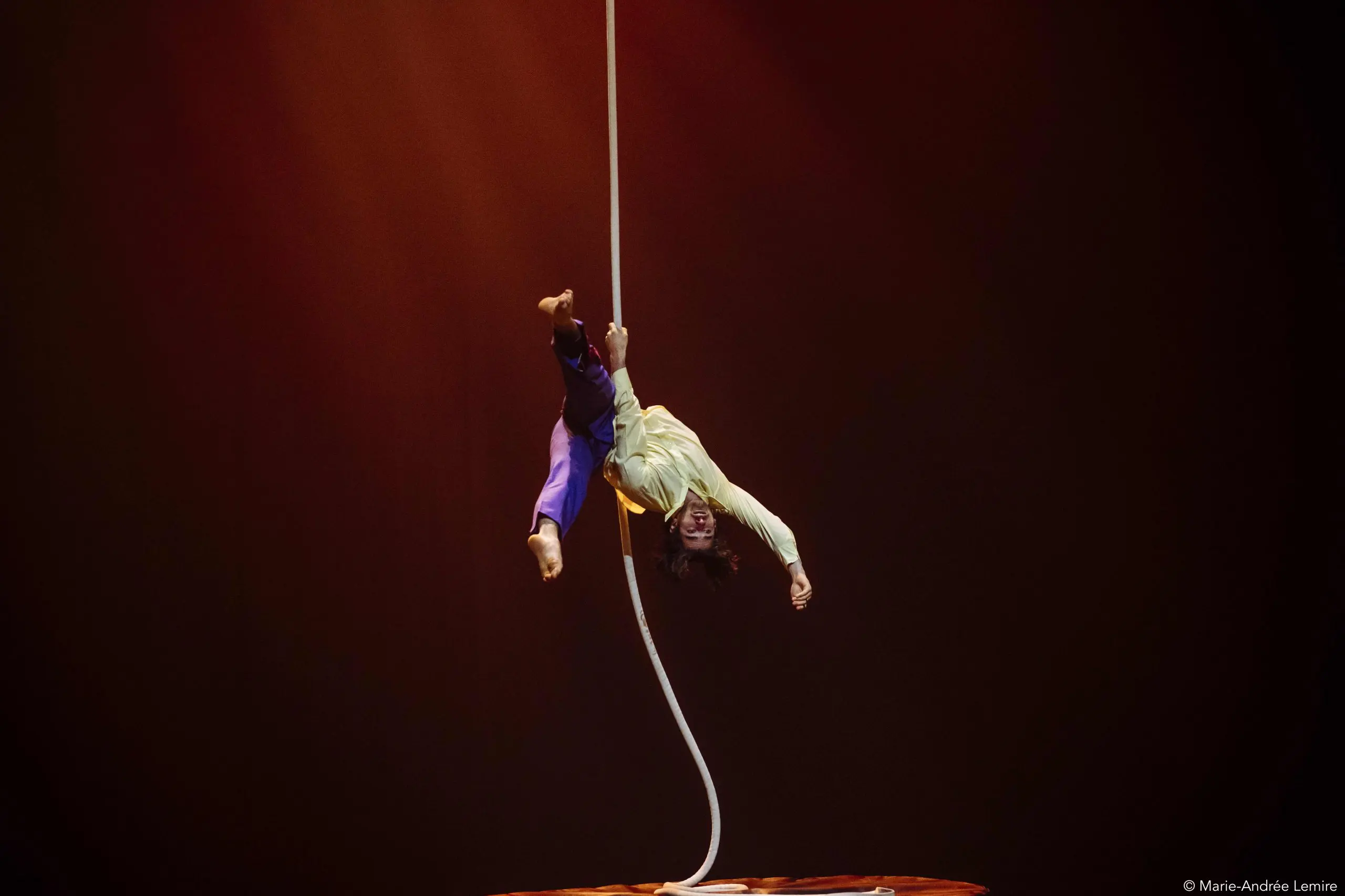 Un artiste de cirque, Dylan Ingwersen, est suspendu à l'envers à une corde aérienne blanche, en plein acte, un bras tendu et les jambes pliées, sur un fond de scène sombre à l'éclairage chaud.