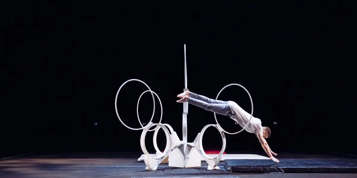 Laurent Racicot, vêtu d'une tenue grise, plonge horizontalement dans une série de cerceaux circulaires blancs disposés sur une scène, dont le fond sombre et les tapis de scène sont visibles.
