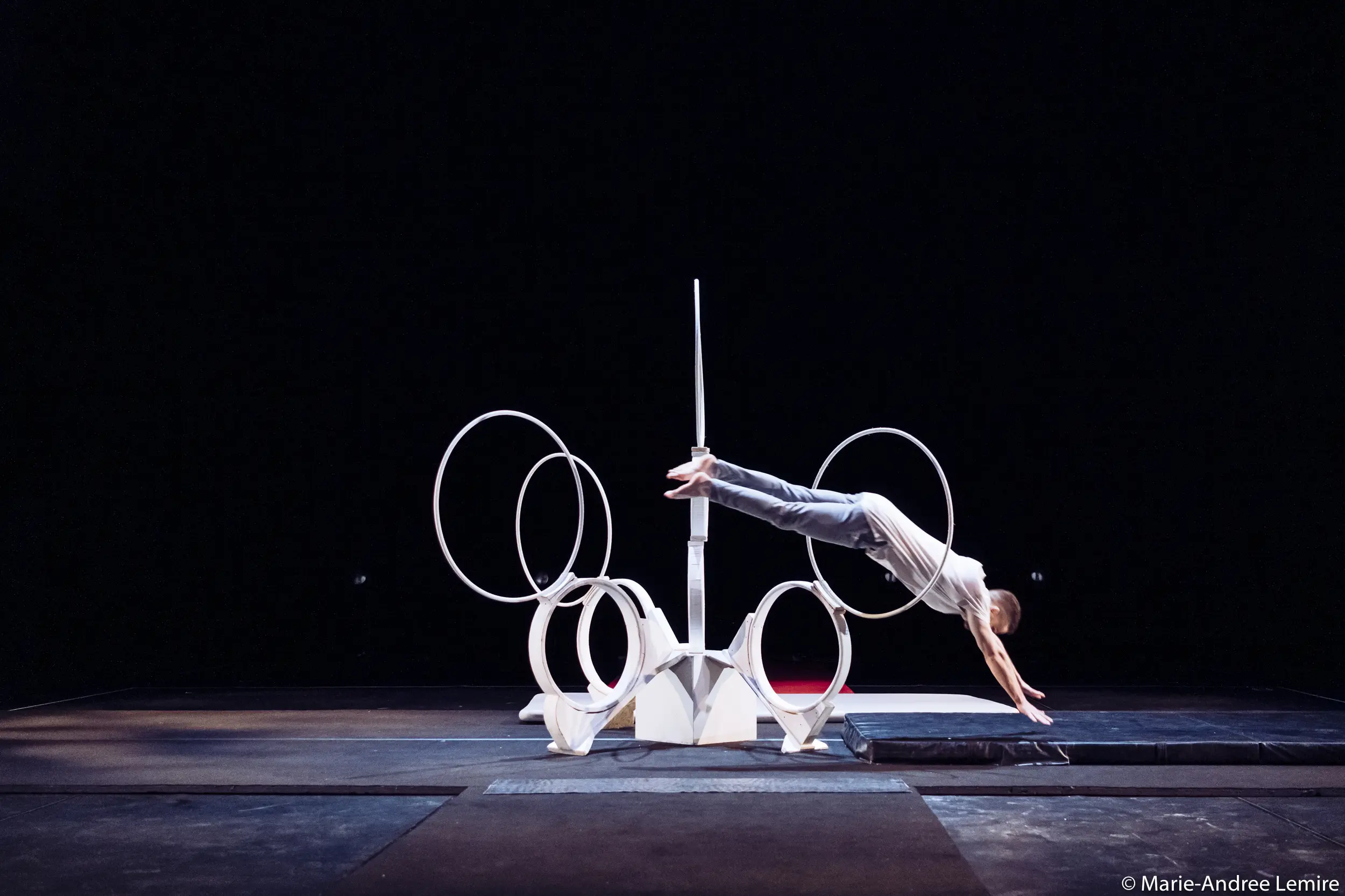 Laurent Racicot, vêtu d'une tenue grise, plonge horizontalement dans une série de cerceaux circulaires blancs disposés sur une scène, dont le fond sombre et les tapis de scène sont visibles.