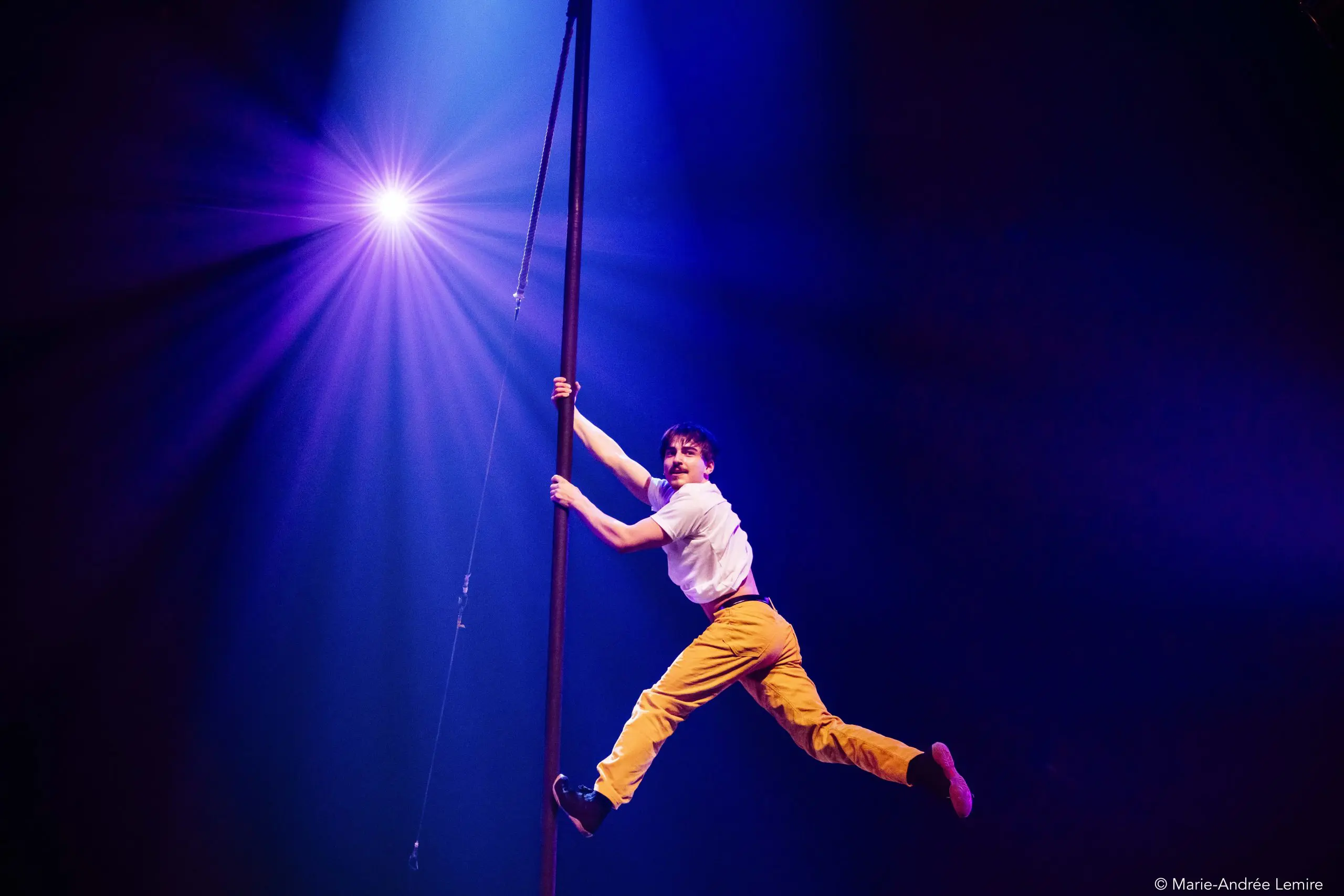 Antoine Brabant, vêtu d'un pantalon jaune et d'une chemise blanche, est suspendu horizontalement à un poteau vertical sur une scène faiblement éclairée par un projecteur violet brillant.