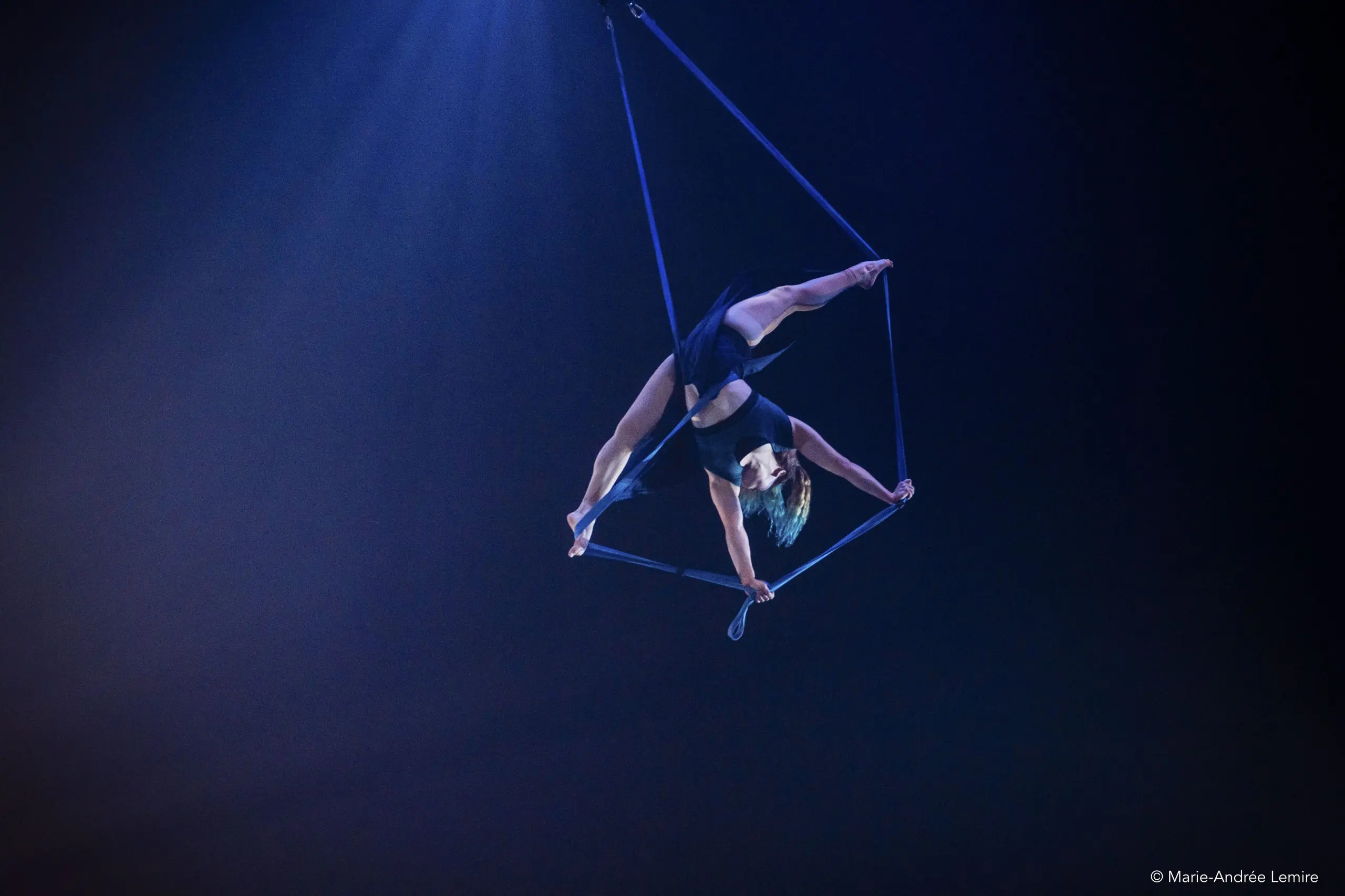 Anja Habegger, vêtue d'une tenue noire, est suspendue la tête en bas en tant qu'artiste aérienne, agrippée à un trapèze suspendu avec ses mains et ses pieds, éclairée par un projecteur bleu sur un fond sombre.