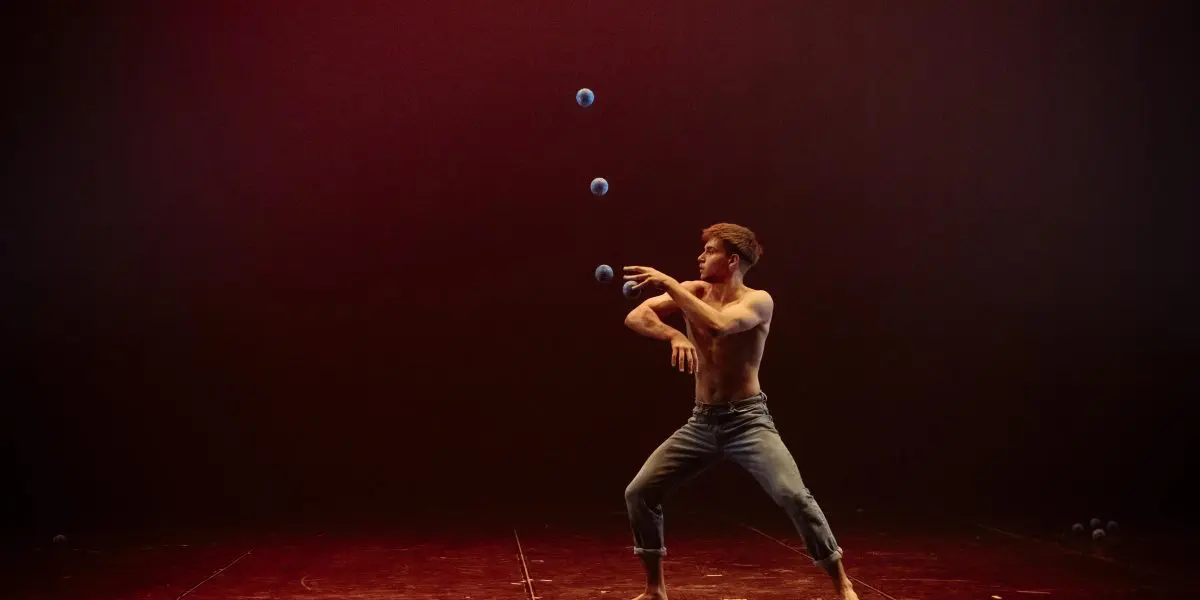 Un homme torse nu portant un jean jongle habilement avec cinq balles bleues sur une scène faiblement éclairée avec un fond brun rougeâtre, rappelant les performances captivantes de Basile Pucek.