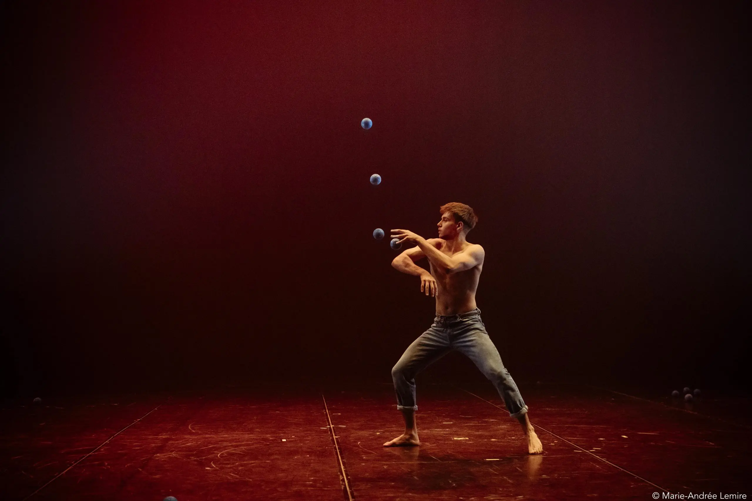 Un homme torse nu portant un jean jongle habilement avec cinq balles bleues sur une scène faiblement éclairée avec un fond brun rougeâtre, rappelant les performances captivantes de Basile Pucek.
