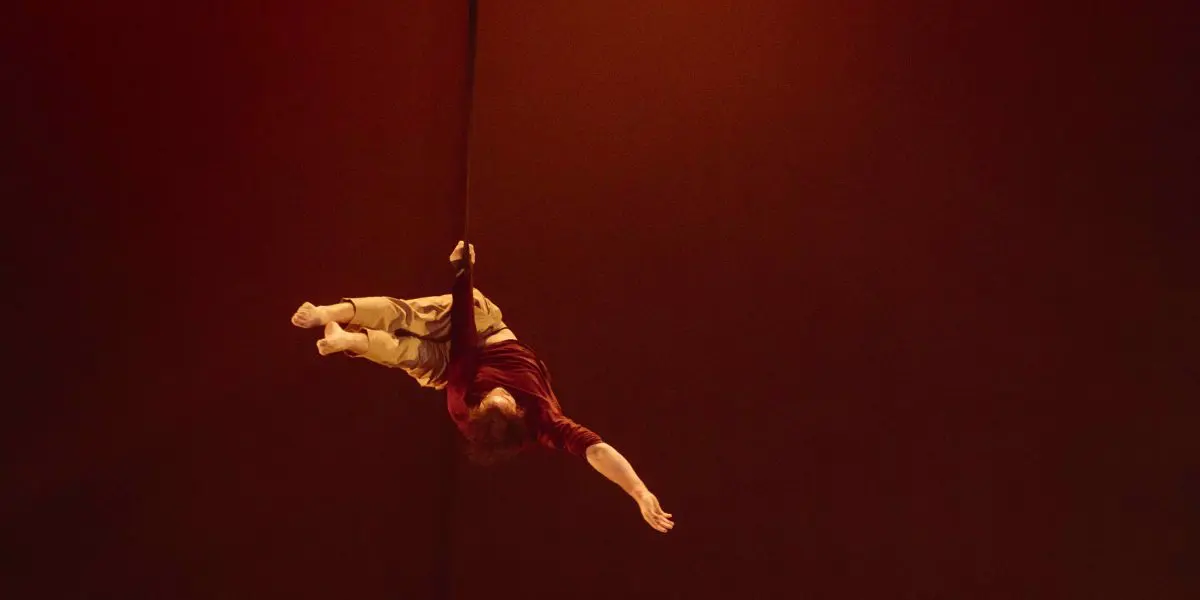 L'artiste de cirque Eduardo De Azevedo Grillo, vêtu d'une chemise rouge et d'un short beige, est suspendu à l'envers à une corde verticale, le bras tendu, sur un fond dramatique brun-orange faiblement éclairé.