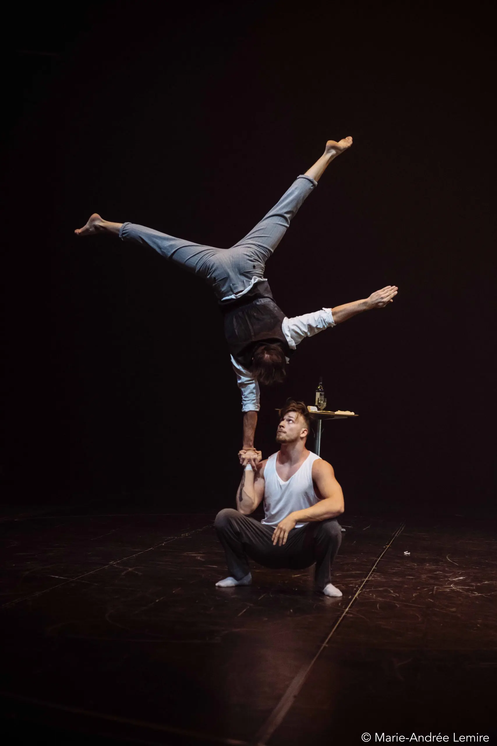 Sur une scène faiblement éclairée, un artiste se tient en équilibre sur une main au dessus de la paume de Carl Löwenborg, assis, les jambes étendues en fente. Une petite table avec une bouteille et un verre est visible à l'arrière-plan.