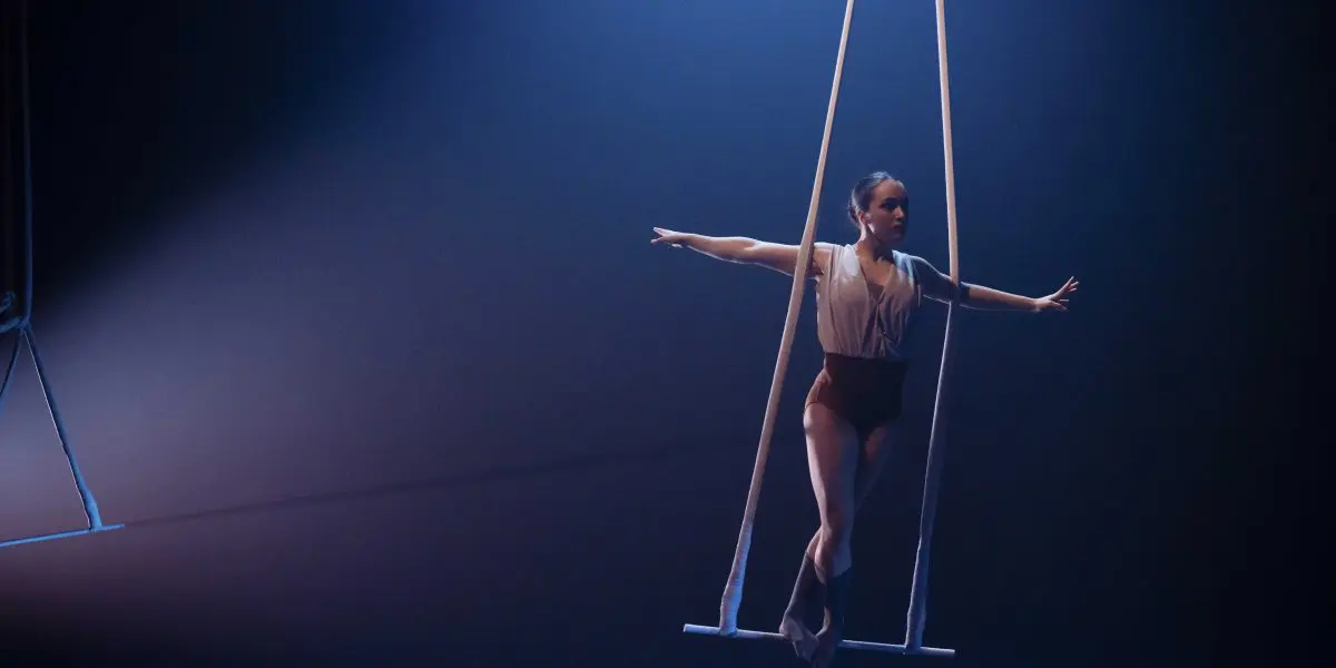 Une voltigeuse, Daphnée Sergerie, se tient gracieusement en équilibre sur un trapèze suspendu dans un théâtre sombre et éclairé, étendant élégamment les bras pendant sa performance.