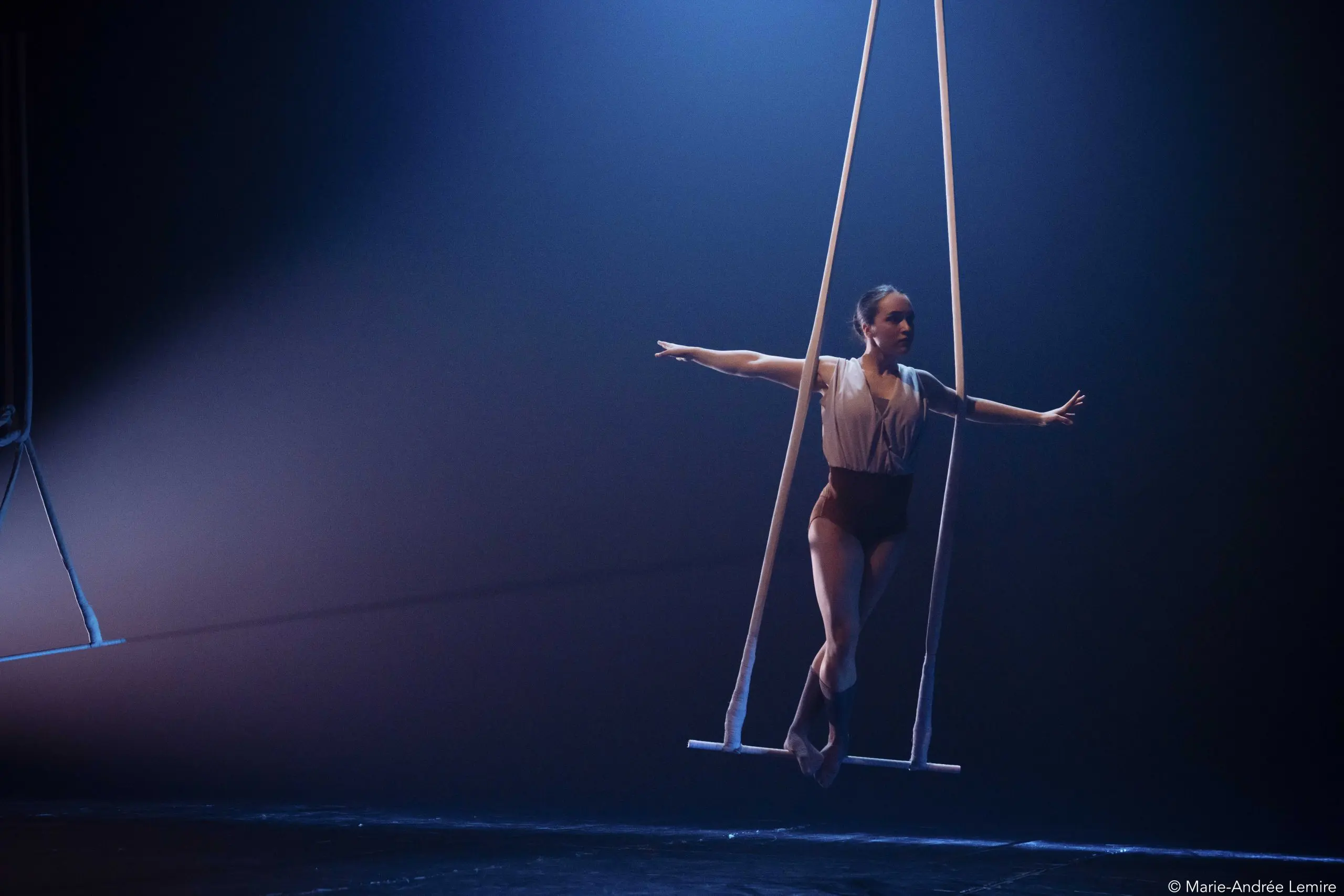 Une voltigeuse, Daphnée Sergerie, se tient gracieusement en équilibre sur un trapèze suspendu dans un théâtre sombre et éclairé, étendant élégamment les bras pendant sa performance.
