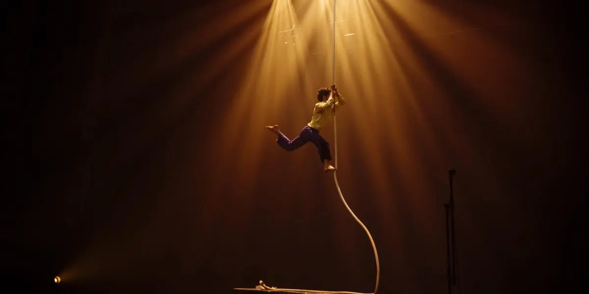 L'artiste Dylan Ingwersen, vêtu d'une chemise verte et d'un pantalon sombre, grimpe sur une corde verticale sur une scène faiblement éclairée, illuminée par des projecteurs dorés spectaculaires provenant d'en haut.