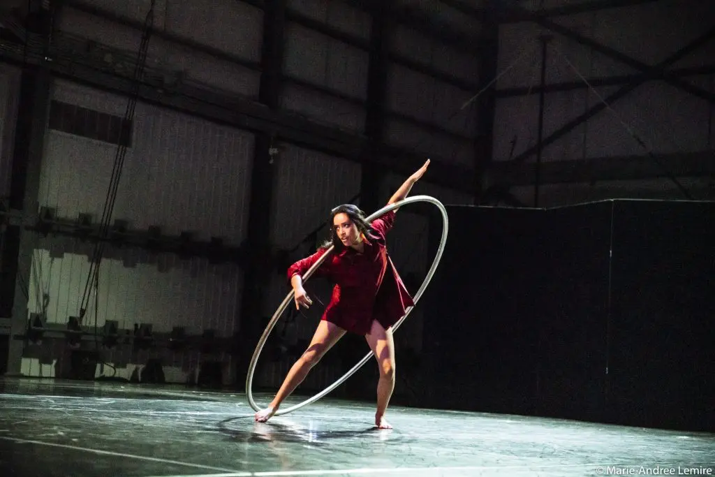 Une femme pieds nus en robe rouge, Andrea Martinez Aguilar, se produit sur scène avec un grand cerceau, en équilibre à l'intérieur de celui-ci, un bras tendu et l'autre touchant le cerceau, sur un fond industriel faiblement éclairé.