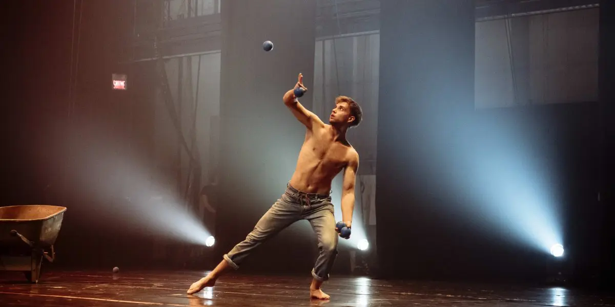 Un artiste en jeans, pieds nus et torse nu - peut-être Basile Pucek - jongle avec trois balles bleues sur une scène faiblement éclairée, des projecteurs dramatiques projetant des ombres tandis qu'une brouette traîne à l'arrière-plan.