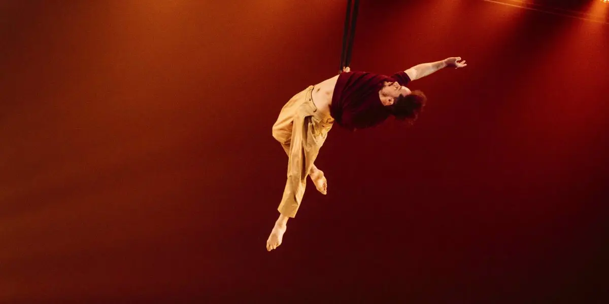 Un artiste aérien, Eduardo De Azevedo Grillo, vêtu d'un pantalon jaune et d'une chemise bordeaux, se suspend gracieusement à une corde sur un fond de scène chaud et éclairé à l'orange, un bras et les deux jambes tendus.