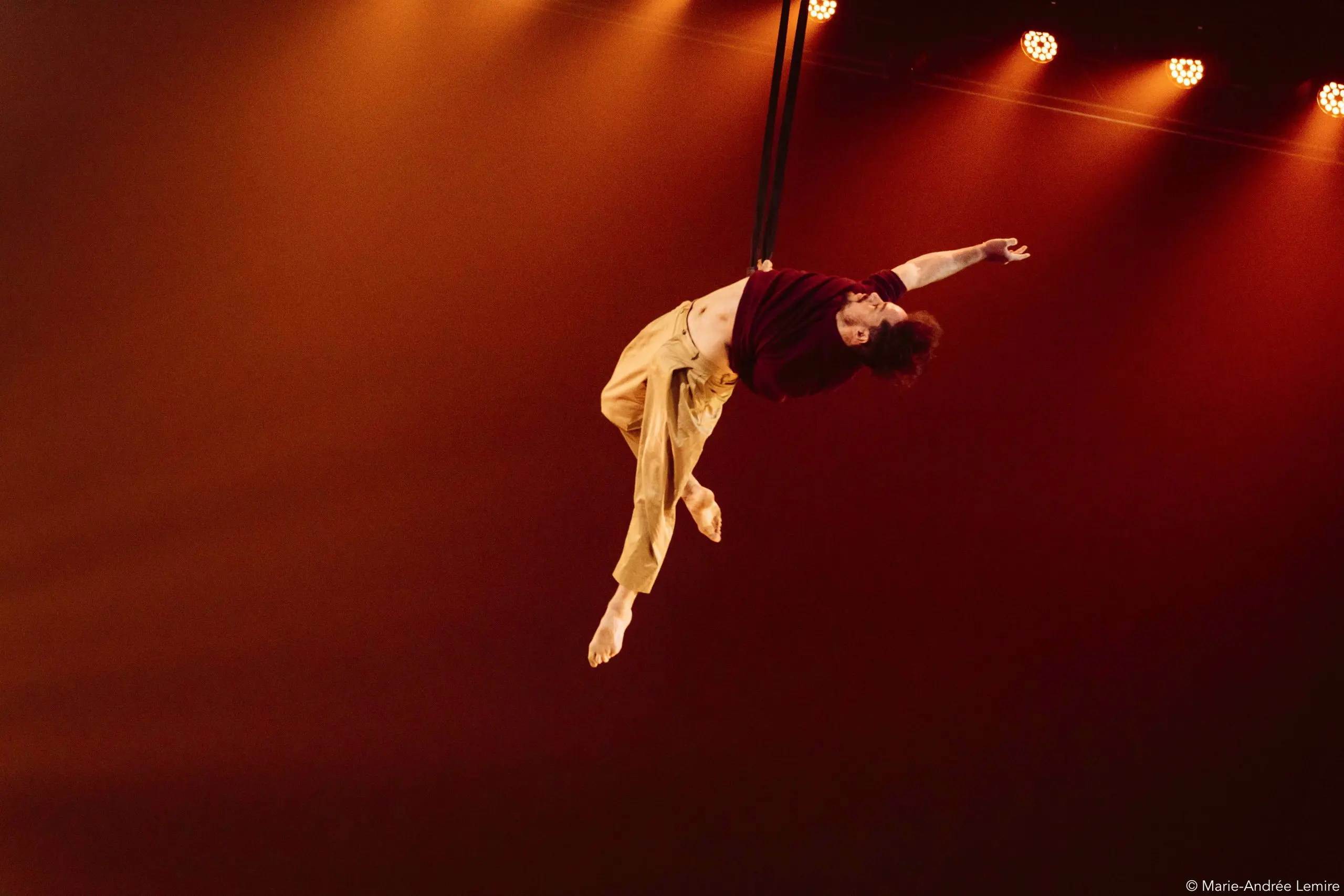 Un artiste aérien, Eduardo De Azevedo Grillo, vêtu d'un pantalon jaune et d'une chemise bordeaux, se suspend gracieusement à une corde sur un fond de scène chaud et éclairé à l'orange, un bras et les deux jambes tendus.