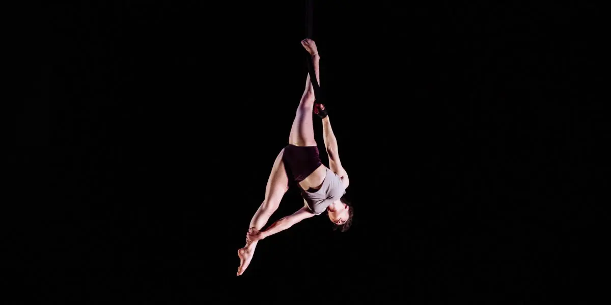 L'acrobate Mathilde Richer suspendue dans les airs sur un fond noir, effectuant une pose acrobatique avec une jambe tendue vers le haut et l'autre pliée, vêtue d'un haut blanc et d'un short noir.