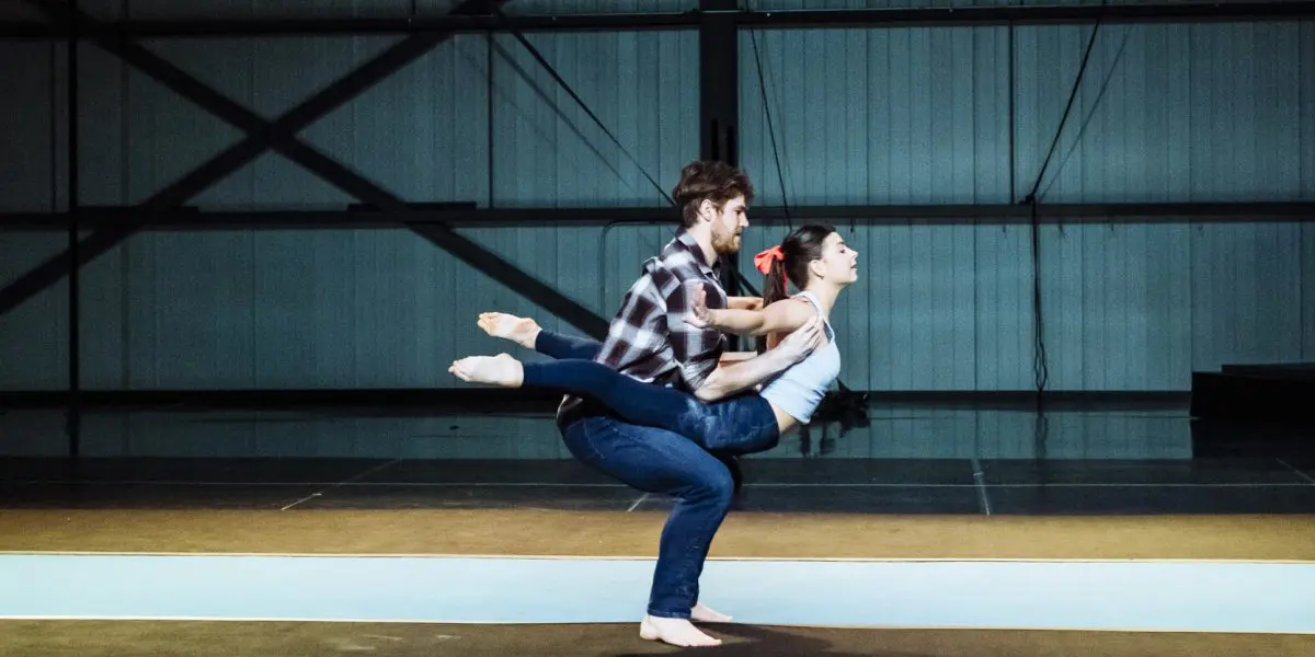 Un homme vêtu d'une chemise à carreaux s'accroupit tandis qu'Ashley Youren, vêtue d'une chemise blanche, se tient en équilibre sur son dos, les bras et une jambe tendus, exécutant un mouvement de danse ou d'acrobatie impressionnant dans un studio de style industriel.
