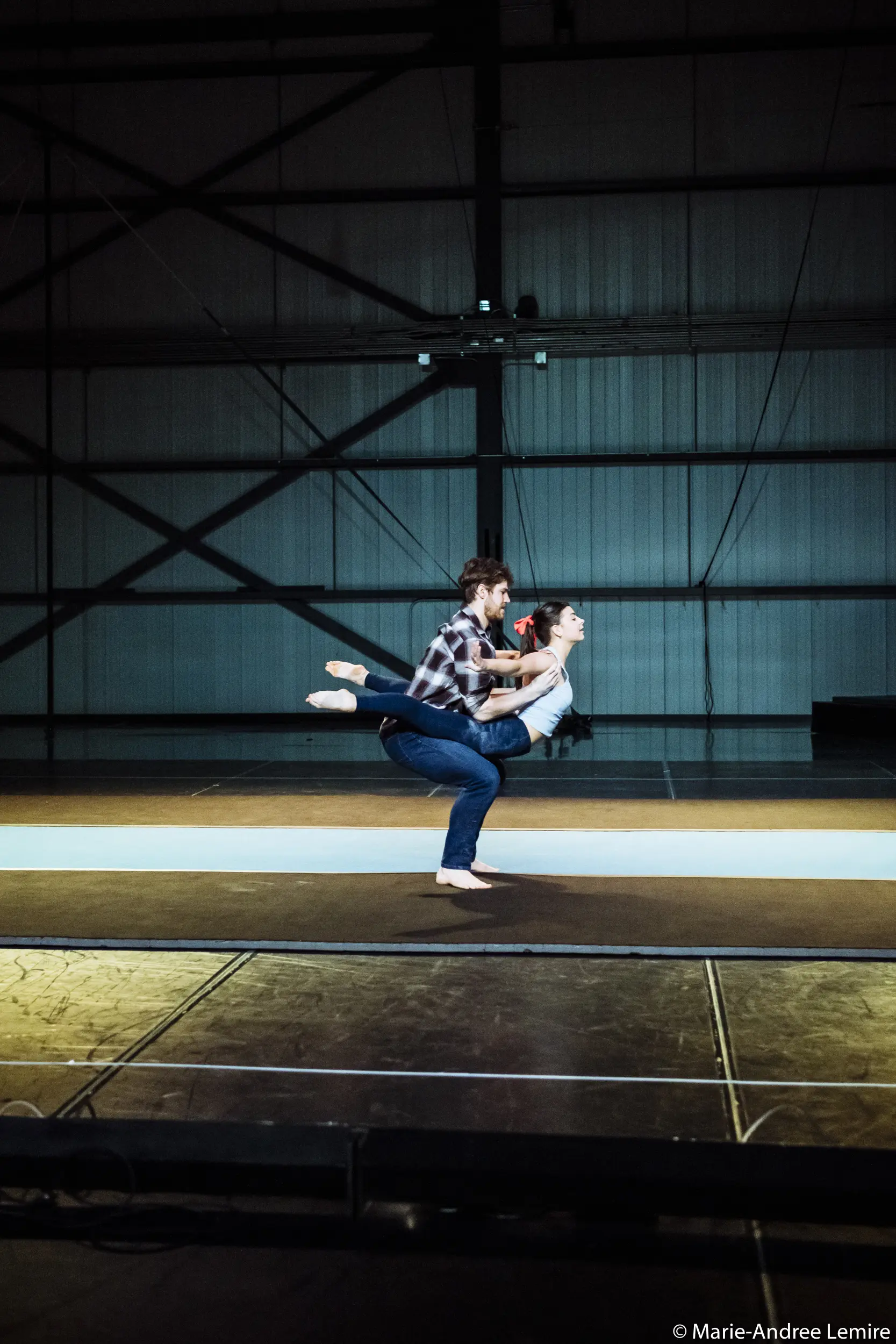 Un homme vêtu d'une chemise à carreaux s'accroupit tandis qu'Ashley Youren, vêtue d'une chemise blanche, se tient en équilibre sur son dos, les bras et une jambe tendus, exécutant un mouvement de danse ou d'acrobatie impressionnant dans un studio de style industriel.