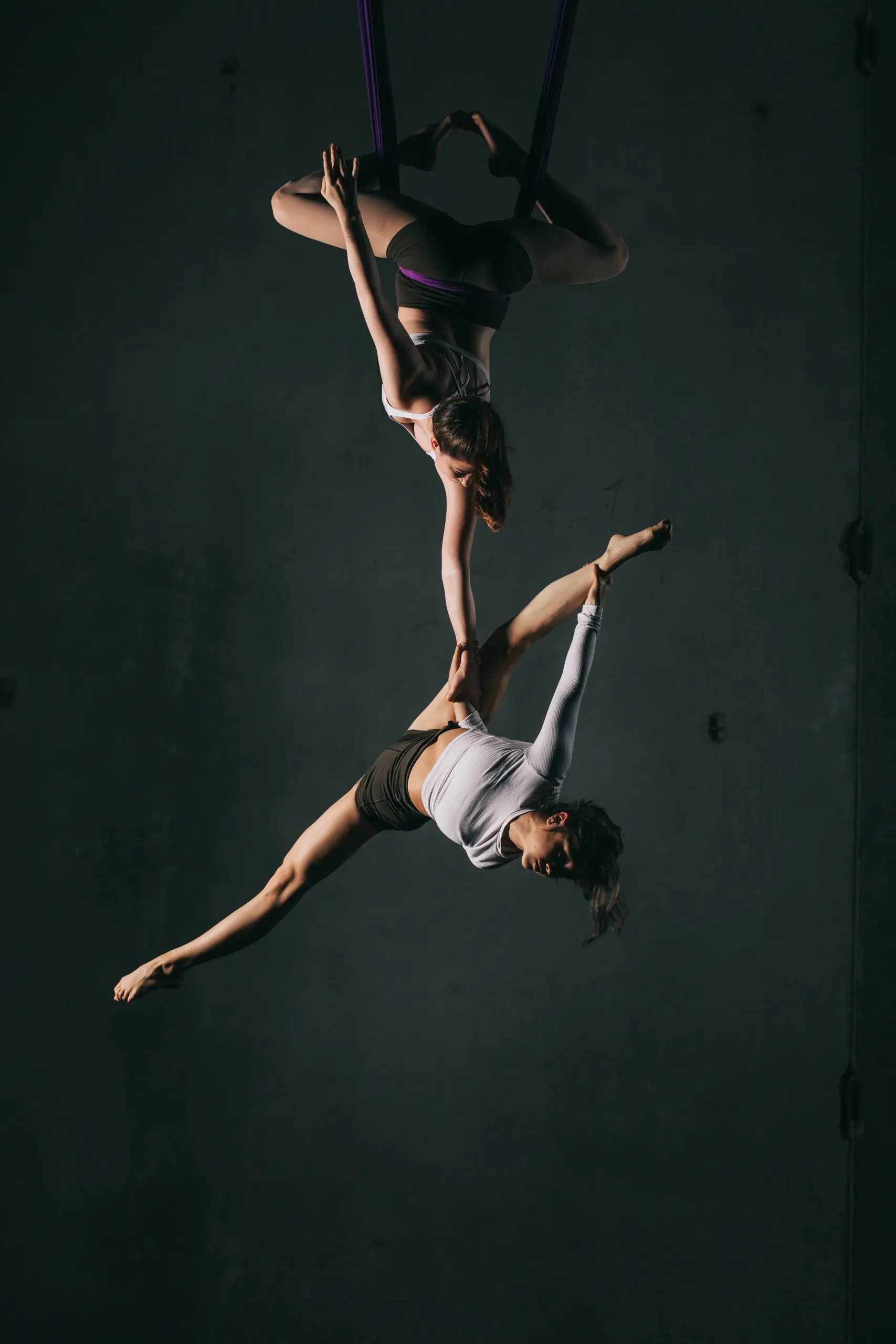 Deux voltigeuses, Sydney Ramirez et Brianna MacDonald, exécutent une pose acrobatique sur des soies ; l'une est suspendue à l'envers en tenant l'autre, qui étend son corps horizontalement en l'air, sur un fond sombre.