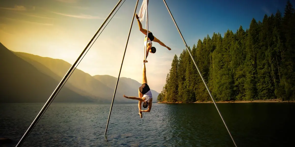 Sydney Ramirez et Brianna MacDonald se produisent sur des soies aériennes au-dessus d'un lac au coucher du soleil, entouré de montagnes et d'arbres boisés, le ciel s'illuminant d'une douce lumière dorée.