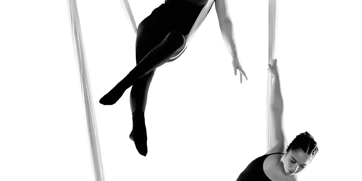 Sydney Ramirez et Brianna MacDonald exécutent une routine aérienne en soie, suspendues dans les airs sur un fond blanc. Elles portent des tenues noires et prennent des poses élégantes en s'agrippant et en se balançant sur le tissu.