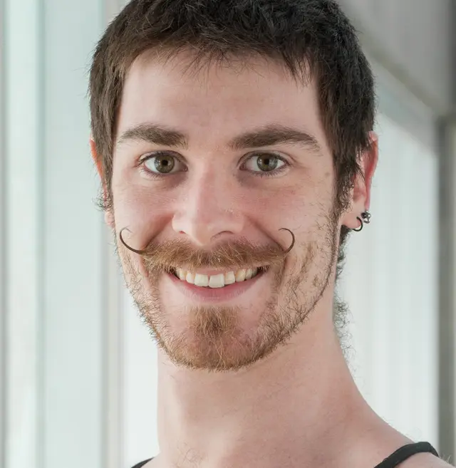 Un jeune homme, Flavien Marciasini, aux cheveux bruns courts, à la moustache frisée et à la barbe légère, sourit à la caméra. Il porte une chemise sans manches et de petites boucles d'oreilles aux deux oreilles. L'arrière-plan lumineux présente de grandes fenêtres.