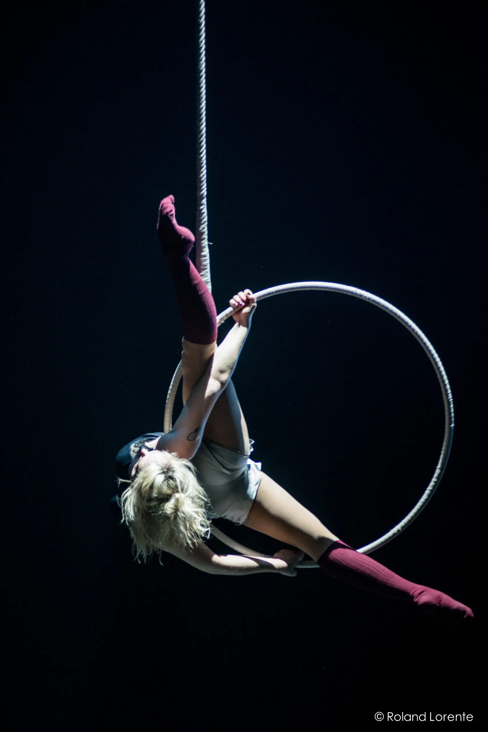 Alyssa Bunce, une artiste aérienne aux cheveux blonds et aux chaussettes rouges à hauteur de cuisse, est suspendue à l'envers à un cerceau, étendant gracieusement ses jambes sur un fond sombre.