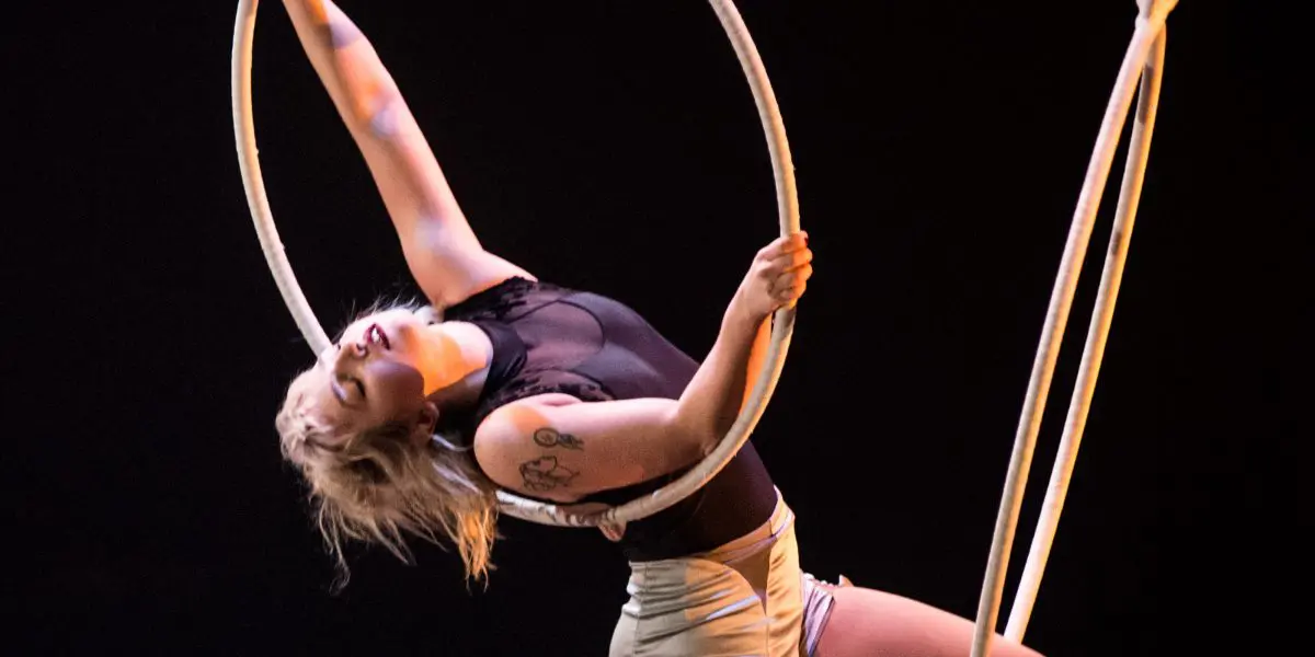 Alyssa Bunce, cheveux blonds courts et tatouages, s'exécute gracieusement sur un cerceau aérien, un bras tendu. Elle porte un haut noir, un short clair et des chaussettes rouges à hauteur des genoux sur un fond sombre.