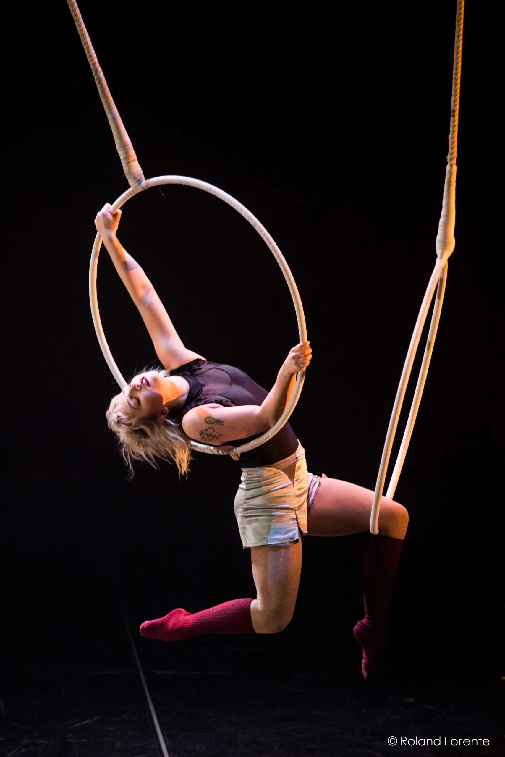 Alyssa Bunce, cheveux blonds courts et tatouages, s'exécute gracieusement sur un cerceau aérien, un bras tendu. Elle porte un haut noir, un short clair et des chaussettes rouges à hauteur des genoux sur un fond sombre.