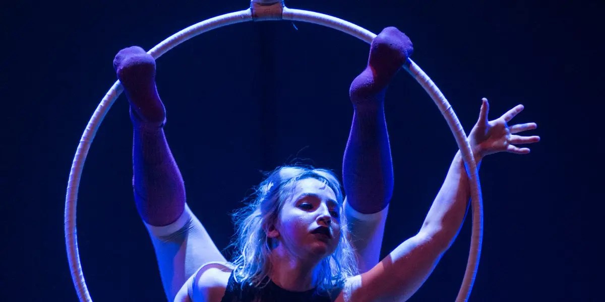 Alyssa Bunce prend une pose acrobatique sur un cerceau suspendu, les jambes tendues vers le haut et les bras tendus, éclairée par un éclairage de scène bleu spectaculaire.