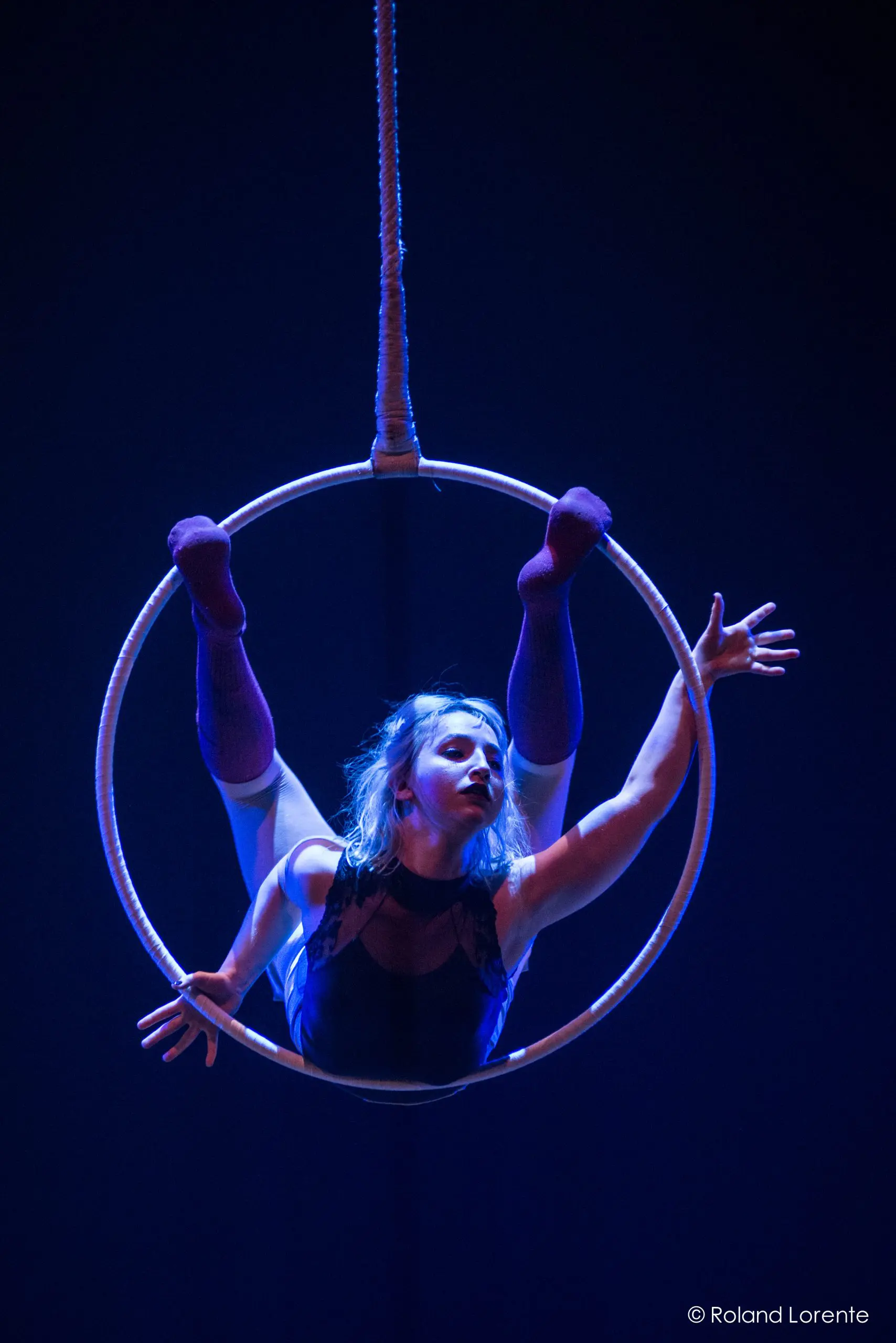 Alyssa Bunce prend une pose acrobatique sur un cerceau suspendu, les jambes tendues vers le haut et les bras tendus, éclairée par un éclairage de scène bleu spectaculaire.